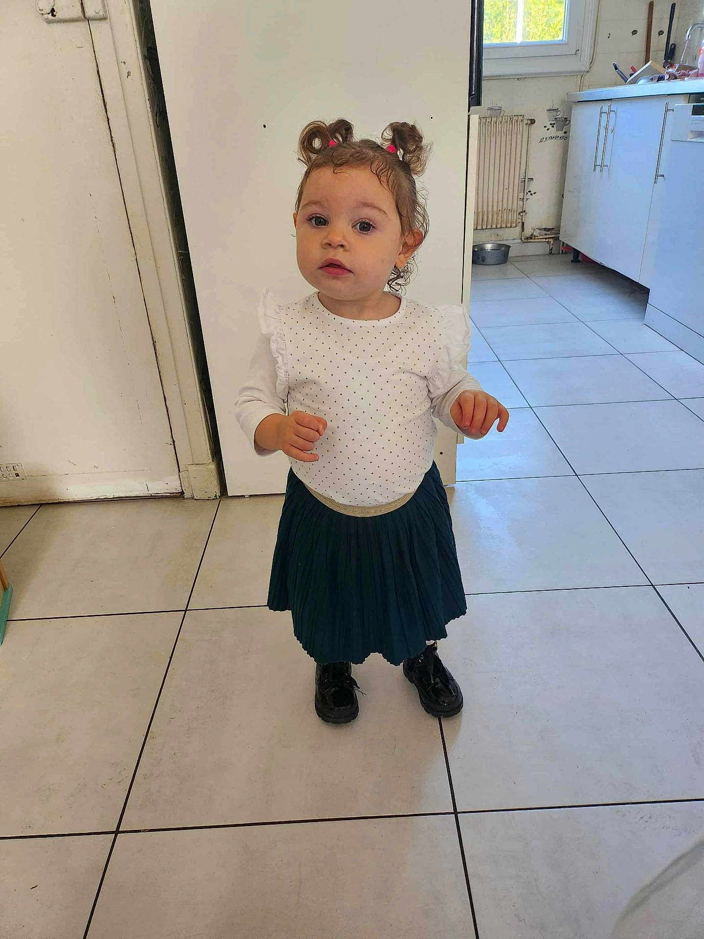 Anna a rejoint le concours — aidez-le/la à gagner de superbes lots ! toddler, child, girl, standing, indoor, kitchen, white_tiles, window, polka_dot_top, pleated_skirt, black_shoes, curly_hair, hair_ties, floor, cabinet, radiator, door, expression, pose, light
