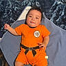 baby, infant, orange_outfit, blanket, soft_texture, happy, smiling, lying_down, indoors, child, cute, portrait, black_hair, short_sleeves, socks, cozy, comfortable, playful, young_child, one_person