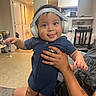 adult, baby, blue_outfit, bottles, carpet, child, dining_area, furniture, headband, headphones, holding, home, indoor, infant, kitchen, parent, photography, portrait, smiling, tongue_out