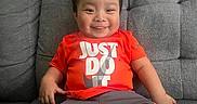 Yael is registered to the contest to win money with this photo: baby, casual_clothing, child, comfortable, couch, cute, face, grey_shorts, happy, home, indoors, orange_shirt, person, playful, short_hair, sitting, smiling, sneakers, white_socks, young_child