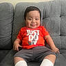 baby, child, smiling, sitting, couch, orange_shirt, grey_shorts, white_socks, sneakers, indoors, happy, person, face, young_child, casual_clothing, short_hair, comfortable, home, cute, playful