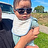 adult_hand, baby, bib, blue_sky, casual, child, clothing, cute, daylight, expression, face, grass, headwear, holding, infant, outdoor, person, portrait, sunglasses, sunny