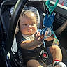 baby, black_clothing, car_interior, car_seat, child, clothing, cute, expression, face, hand, indoor, infant, reaching, red_bracelet, seatbelt, sock, striped_cushion, sunlight, toy, window