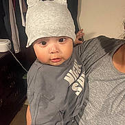Yael joined the competition — help win amazing prizes! baby, child, hat, gray_clothing, indoor, person, adult, holding, face, cute, eyes, expression, clothes, furniture, wall, human, portrait, headwear, sleeve, skin