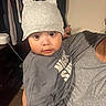 baby, child, hat, gray_clothing, indoor, person, adult, holding, face, cute, eyes, expression, clothes, furniture, wall, human, portrait, headwear, sleeve, skin