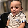 toddler, child, smiling, polo_shirt, beige_clothing, sitting, black_chair, indoor, face, happy, short_hair, person, young_child, casual_clothing, portrait, cute, baby, seated, background_blur, expression