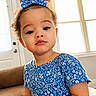 toddler, child, curly_hair, bow, blue_dress, floral_pattern, indoor, couch, window, natural_light, portrait, person, cute, sitting, young_child, face, expression, home, casual_clothing, daylight