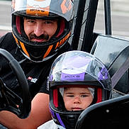 Kaïden participe au concours pour gagner de l'argent avec cette photo : child, adult, helmet, go_kart, racing, seatbelt, race_track, vehicle, safety_gear, outdoor, person, sport, family, transportation, helmet_visors, black_clothing, purple_helmet, orange_helmet, driving, fun