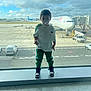 child, pacifier, airport, window, airplane, air_france, tarmac, vehicle, cloudy_sky, sneakers, green_pants, white_shirt, glass, standing, boy, travel, daylight, reflection, casual_clothing, outdoor_view
