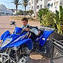 child, boy, quad_bike, blue_vehicle, outdoor, sunny, palm_trees, white_buildings, playful, smiling, shorts, sandals, sidewalk, urban, plants, greenery, daytime, recreation, motor_vehicle, adventure