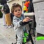 child, toddler, bicycle, training_wheels, denim_jacket, curly_hair, outdoor, party, balloons, adults, wooden_floor, casual_clothing, sneakers, fence, window, curious_expression, green_bike, celebration, people, daylight
