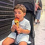 child, stroller, ice_cream, cone, shopping_mall, floor, people, conversation, casual_clothing, shorts, tshirt, footwear, indoor, black_stroller, happy, snack, summer, cute, young_child, sitting