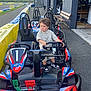 child, go_kart, karting, outdoor, track, racing, vehicle, steering_wheel, bench, barrier, asphalt, helmet_absent, curly_hair, sneakers, clothing, number_one, building, daylight, road, person