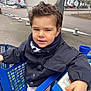 child, boy, curly_hair, jacket, scarf, shopping_cart, outdoor, urban, sidewalk, cars, trees, winter, street, hand, seat, clothing, person, portrait, expression, playful