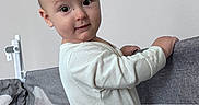 Daniel is registered to the contest to win money with this photo: baby, child, infant, onesie, playpen, indoor, portrait, curious, smiling, standing, face, head, hands, white_clothing, light, wall, room, young, cute, looking