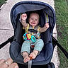 baby, stroller, child, smile, happy, toy, outdoor, sidewalk, grass, hand, person, clothing, footwear, seat, safety_harness, infant, playful, portrait, casual, parent