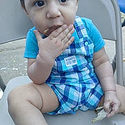 Armando is registered to the contest to win money with this photo: baby, child, toddler, blue_clothing, plaid, romper, sitting, chair, food, eating, hand, face, outdoor, curious, cute, expression, skin, legs, person, young