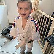 Ismaël participe au concours pour gagner de l'argent avec cette photo : toddler, child, martial_arts_uniform, karate_gi, white_belt, toy_car, indoor, floor_tiles, baby_gate, exercise_equipment, window, curtains, smiling, happy, short_hair, sock, standing, person, home, casual