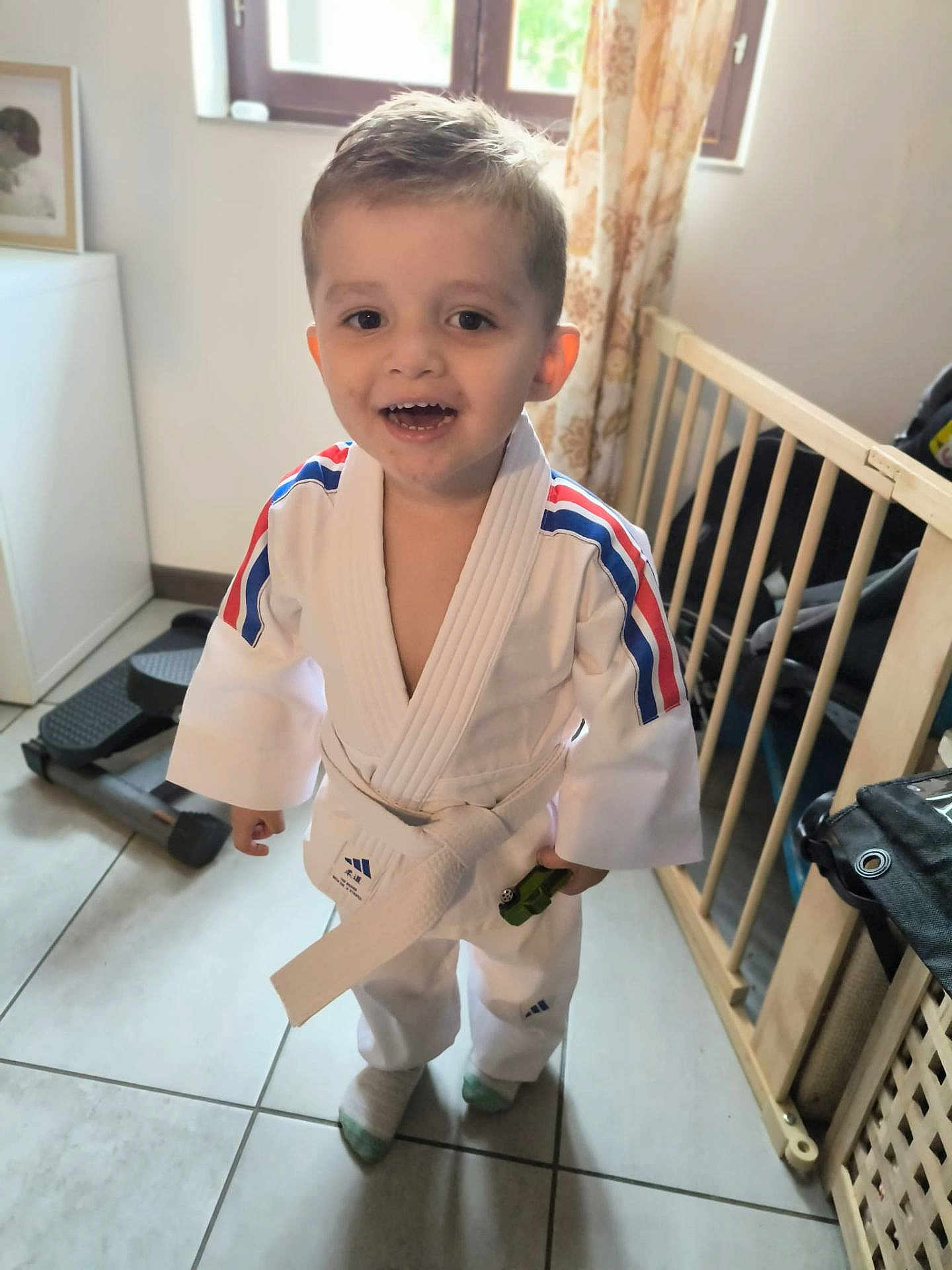 Ismaël participe au concours pour gagner de l'argent avec cette photo : toddler, child, martial_arts_uniform, karate_gi, white_belt, toy_car, indoor, floor_tiles, baby_gate, exercise_equipment, window, curtains, smiling, happy, short_hair, sock, standing, person, home, casual