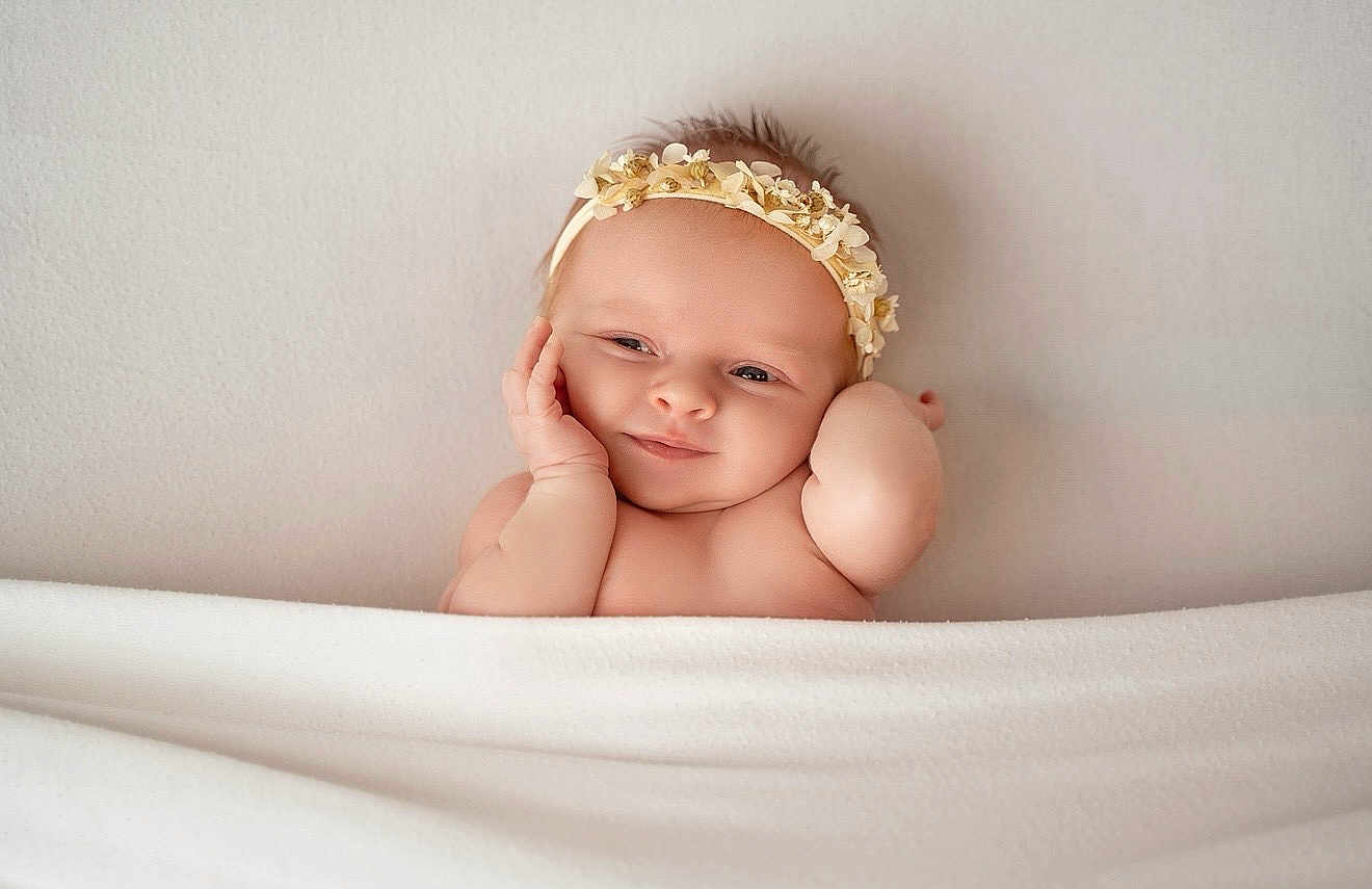 Eliza is registered to the contest to win money with this photo: baby, infant, headband, flower_crown, smile, blanket, skin, face, cheeks, hands, newborn, cute, portrait, resting, soft_texture, neutral_background, peaceful, indoors, toddler, young_child