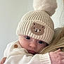 Eliza is registered to the contest to win money with this photo: baby, infant, knit_hat, pom_pom, bear_patch, cream_sweater, wooden_button, face, eyes, hand, cuddling, person, close_up, indoor, soft_lighting, warm_clothing, cute, child, portrait, cozy