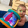 Josiah is registered to the contest to win money with this photo: baby, diaper, toy, rug, indoor, face_paint, glowing, colorful, curious, couch, carpet, child, infant, playtime, blue, orange, pink, green, living_room, cute