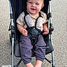 baby, stroller, child, smiling, outdoor, pavement, barefoot, clothing, safety_harness, footrest, person, infant, cute, happy, seat, casual, young_child, playful, daylight, portrait