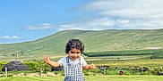 Aydin is registered to the contest to win money with this photo: child, toddler, overalls, gingham, walking, grass, meadow, countryside, hill, blue_sky, clouds, landscape, smile, portrait, outdoors, shoes, fence, vacation, greenery, sunny_day