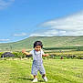 child, toddler, overalls, gingham, walking, grass, meadow, countryside, hill, blue_sky, clouds, landscape, smile, portrait, outdoors, shoes, fence, vacation, greenery, sunny_day