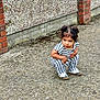 Aydin is registered to the contest to win money with this photo: child, toddler, squatting, striped_outfit, hair_buns, concrete, pavement, brick_pillar, pebble_wall, sneakers, outdoor, curious_expression, portrait, candid, young_child, sidewalk, urban, small_person, daylight, playful