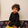 Aydin is registered to the contest to win money with this photo: child, toddler, curly_hair, black_outfit, sitting, stool, plastic_stool, portrait, indoor, white_background, patterned_wallpaper, hand, eyes, looking_at_camera, cute, studio, small_hands, neutral_expression, footwear, person