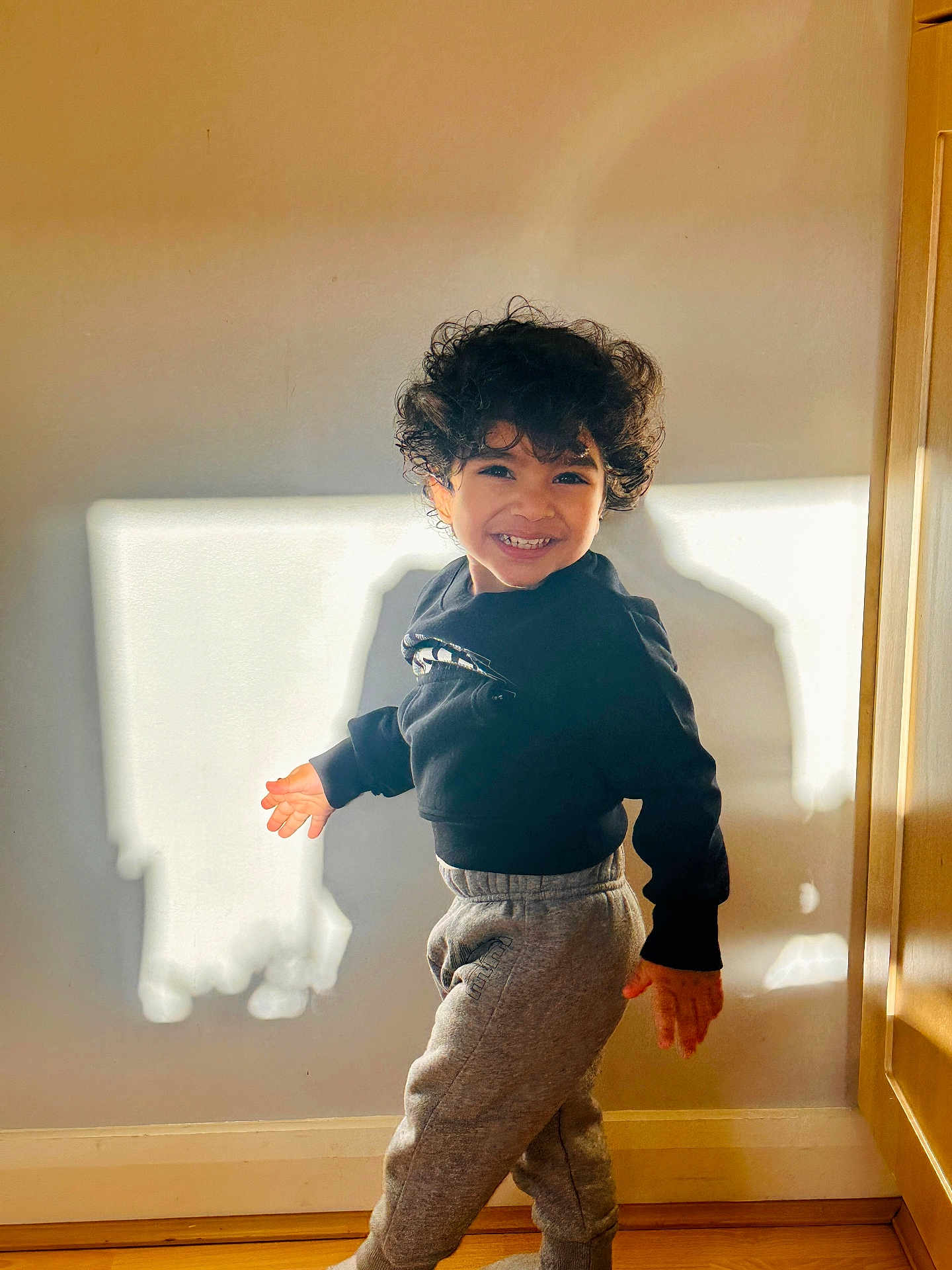 Aydin is registered to the contest to win money with this photo: child, smiling, curly_hair, sweatshirt, sweatpants, indoor, sunlight, shadow, wall, wooden_floor, candid, portrait, playful, happy, teeth, standing, home_interior, natural_light, doorframe, young_kid