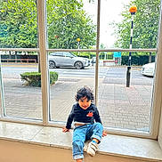 Aydin joined the competition — help win amazing prizes! child, toddler, window, windowsill, street, car, tree, lamppost, sidewalk, pavement, smile, jeans, sweatshirt, shoes, indoor, glass_window, bench, portrait, curb, playful