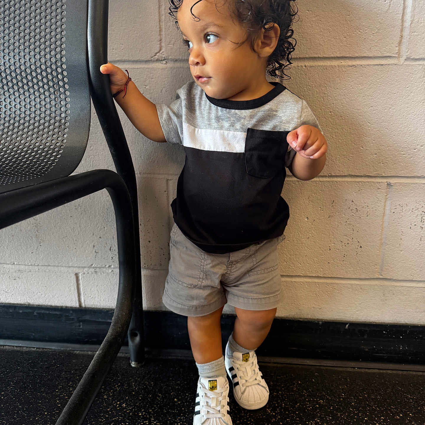 Lahiam is registered to the contest to win money with this photo: black_and_gray, brick_wall, casual_clothing, child, curious_expression, curly_hair, floor, footwear, holding, indoor, looking_away, metal_chair, person, shorts, small_child, sneakers, standing, toddler, tshirt, young_child