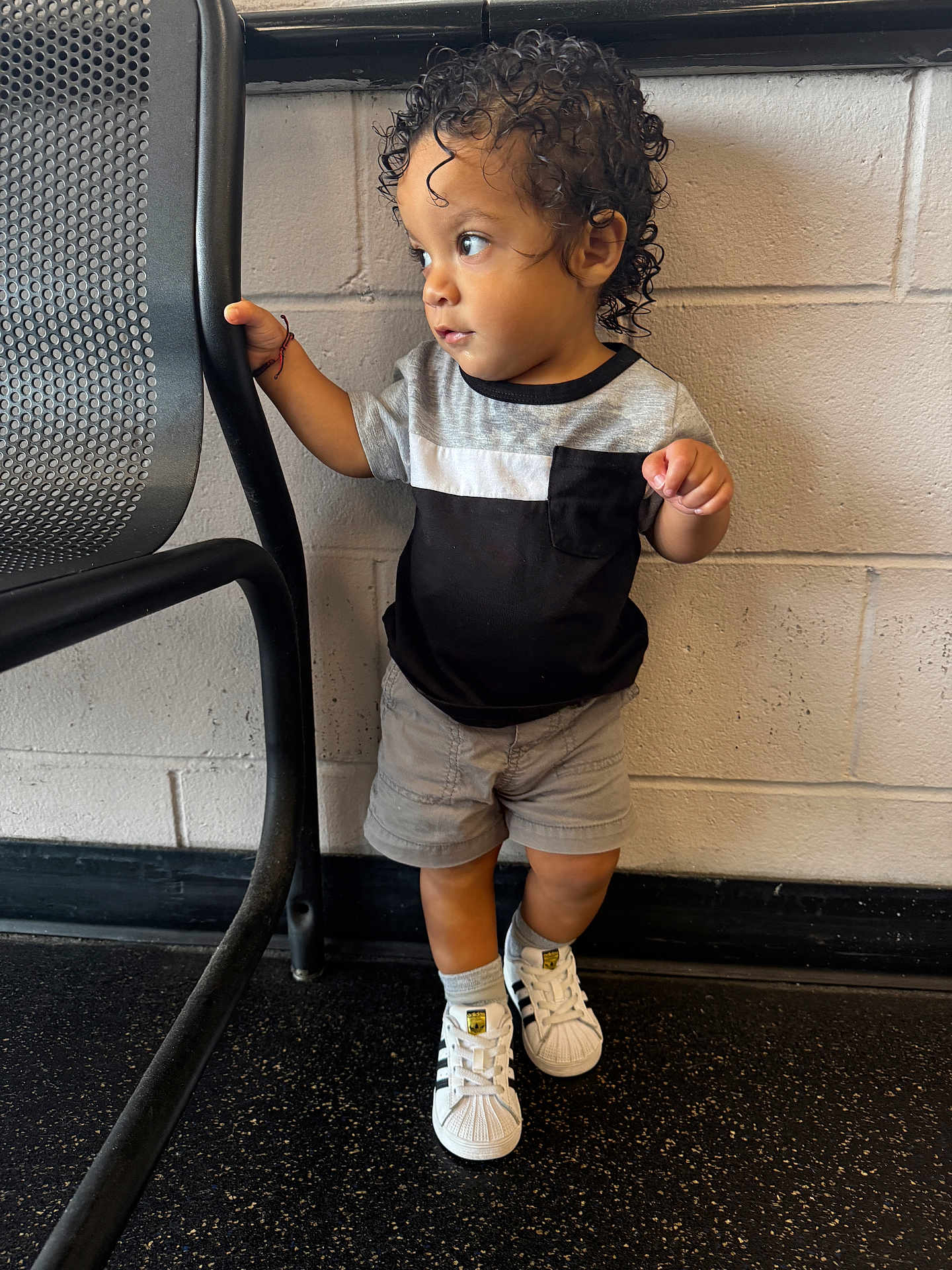 Lahiam is registered to the contest to win money with this photo: toddler, child, curly_hair, standing, sneakers, shorts, tshirt, black_and_gray, metal_chair, brick_wall, floor, indoor, casual_clothing, curious_expression, footwear, small_child, holding, looking_away, person, young_child