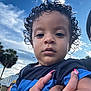Lahiam is registered to the contest to win money with this photo: child, toddler, curly_hair, outdoor, sky, clouds, palm_tree, hand, nails, person, blue_shirt, face, portrait, daylight, nature, holding, looking, casual_clothing, expression, closeup