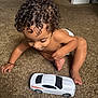 child, toddler, toy_car, curly_hair, carpet, indoor, diaper, playing, crawling, brown_skin, cute, baby, floor, car, foot, hand, room, door, home