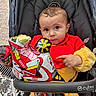 baby, child, stroller, balloon, confetti, red_clothing, yellow_clothing, outdoor, curious_expression, person, face, hand, toy, seat, fabric, wheel, wheelchair, plush, cushion, playful