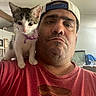 man, kitten, cat, indoor, shoulder, cap, collar, bell, red_tshirt, facial_expression, furniture, living_room, curious, pet, animal, portrait, casual, headwear, person, closeup