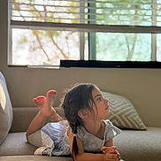Eva is registered to the contest to win money with this photo: child, girl, braids, couch, indoor, window, blinds, pillow, sunlight, shadow, relaxing, portrait, side_view, casual_clothing, home, comfortable, young, resting, daylight, thinking