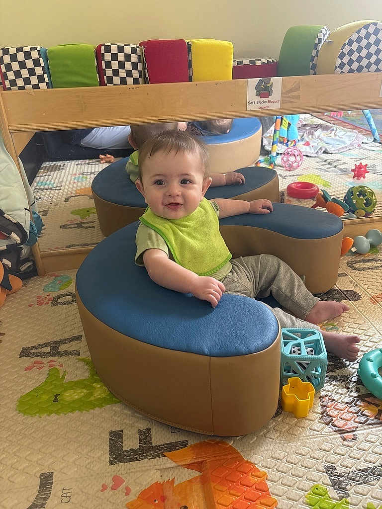 Ezra joined the competition — help win amazing prizes! baby, child, green_bib, blue_cushion, tan_cushion, toys, colorful, playmat, mirror, reflection, indoor, smiling, seated, soft_furniture, playroom, cute, happy, infant, floor_mat, patterned_mat