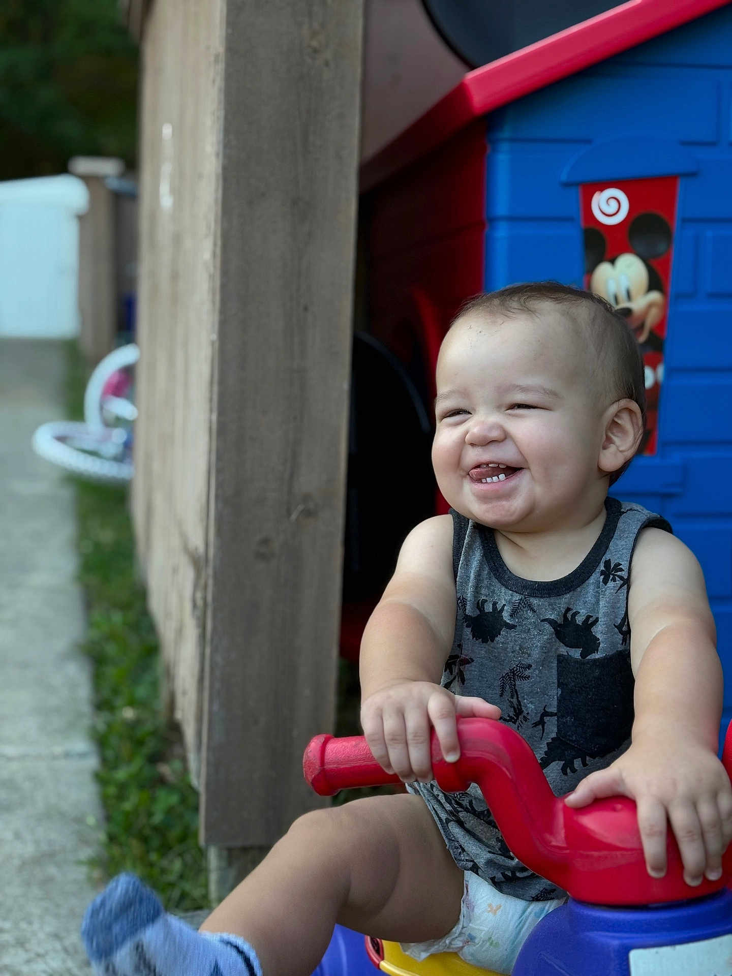 Amor is registered to the contest to win money with this photo: toddler, child, smiling, tricycle, toy, outdoor, playhouse, mickey_mouse, plastic, fence, grass, sidewalk, diaper, tank_top, happy, baby, person, cute, playtime, summer