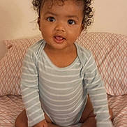 Semaj joined the competition — help win amazing prizes! baby, child, curly_hair, striped_onesie, sitting, bedspread, patterned_bedspread, indoors, cute, portrait, infant, person, smiling, happy, feet, hands, wall, young, looking_at_camera, expression