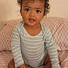 baby, child, curly_hair, striped_onesie, sitting, bedspread, patterned_bedspread, indoors, cute, portrait, infant, person, smiling, happy, feet, hands, wall, young, looking_at_camera, expression