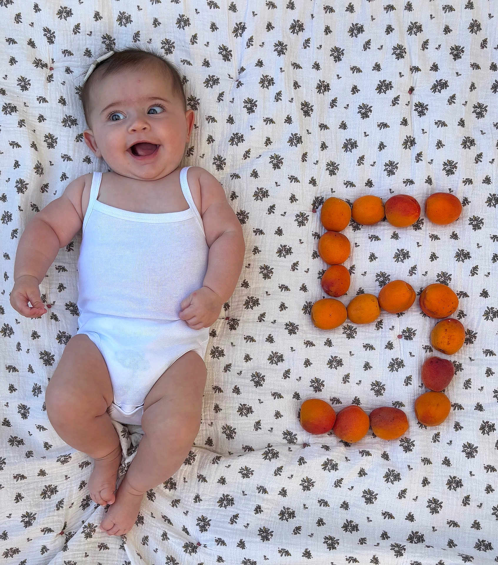 Adèle a rejoint le concours — aidez-le/la à gagner de superbes lots ! baby, infant, smiling, white_onesie, blanket, floral_pattern, apricots, fruit, number_five, outdoor, cute, happy, lying_down, skin, headband, chubby, barefoot, natural_light, playful, portrait