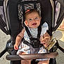 baby, smiling, stroller, child, toy, sunlight, happy, cute, infant, clothing, hair_clip, seatbelt, outdoor, sitting, person, portrait, baby_dress, footwear, playful, daylight