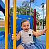 Zayden is registered to the contest to win money with this photo: toddler, child, smiling, playground, outdoors, play_equipment, yellow_bars, blue_platform, striped_shorts, white_shirt, sitting, grinning, happy, face, portrait, sunny, trees, sky, park, recreation