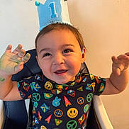 Zayden is registered to the contest to win money with this photo: baby, child, toddler, birthday, crown, party_hat, smiling, happy, portrait, sitting, high_chair, indoors, colorful, clothing, patterned_shirt, hands_up, celebration, face, dark_hair, photography