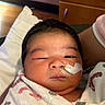 newborn, baby, infant, face, portrait, sleepy, hospital, blanket, swaddled, medical_tube, tape, cheek, dark_hair, eyes, nose, mouth, bed, pillow, adult_arm, indoor