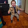 toddler, child, indoor, wooden_floor, plaid_jacket, boots, pants, tongue_out, smiling, standing, dresser, door, window, light_switch, home, diapers_box, playful, person, flooring, casual_clothing